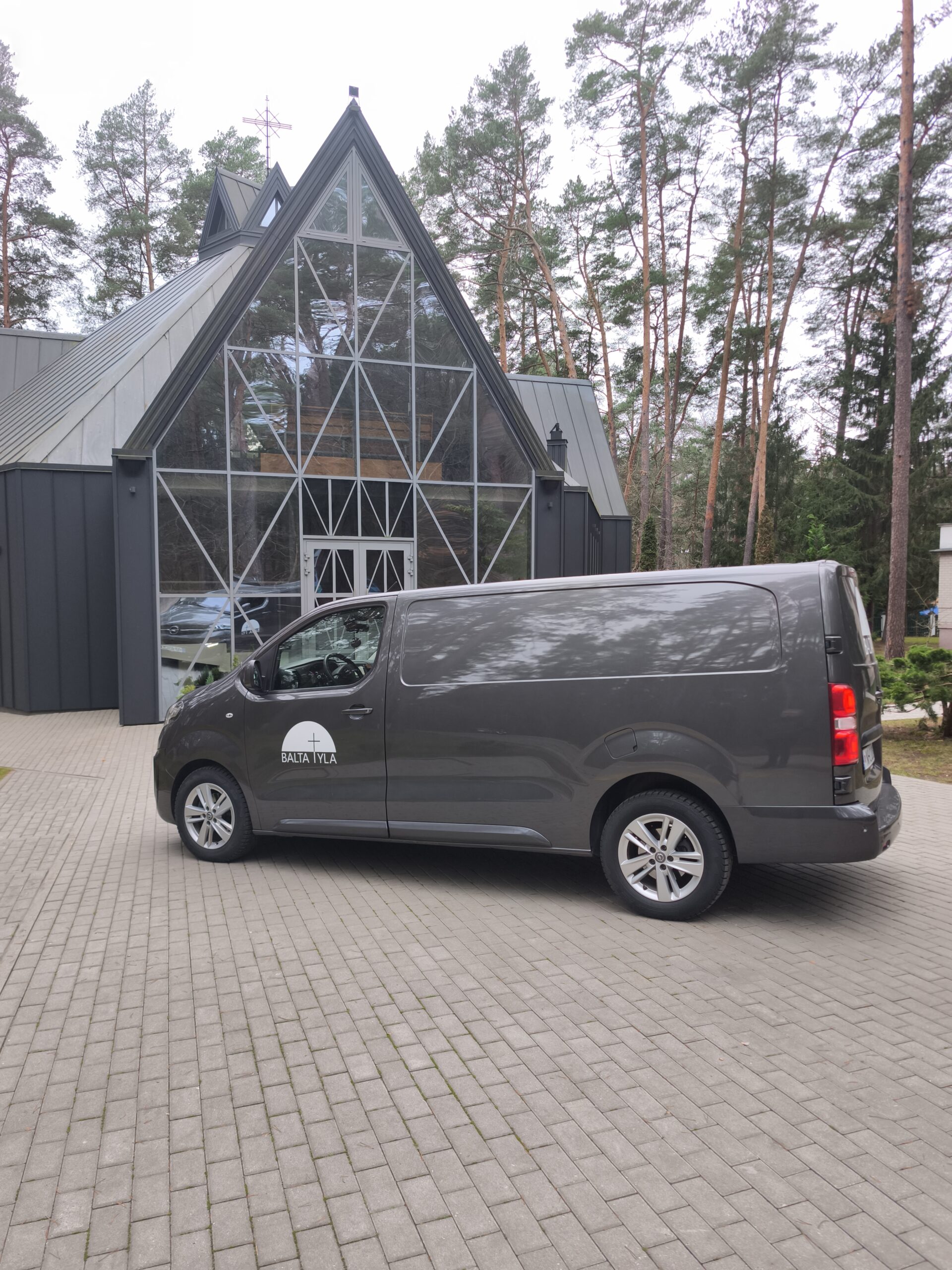 Balta tyla objektas – pilkas mikroautobusas prie modernaus A formos namo su stikline vitrina miško aplinkoje.