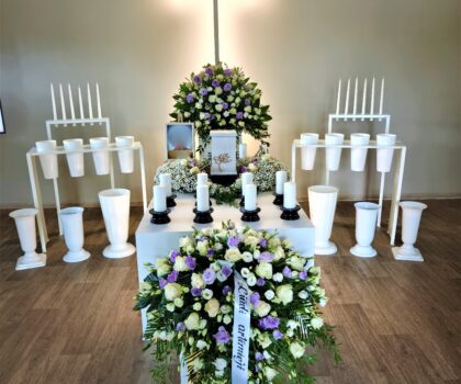 Atsisveikinimo ceremonija salėje su kryžiumi, gėlių kompozicijomis ir karstu centre.