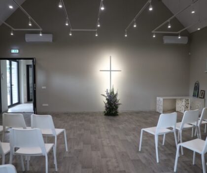 Minimalistinė ceremonijų salė su kryžiumi ant sienos, baltomis kėdėmis ir centrine dekoratyvine augalo kompozicija